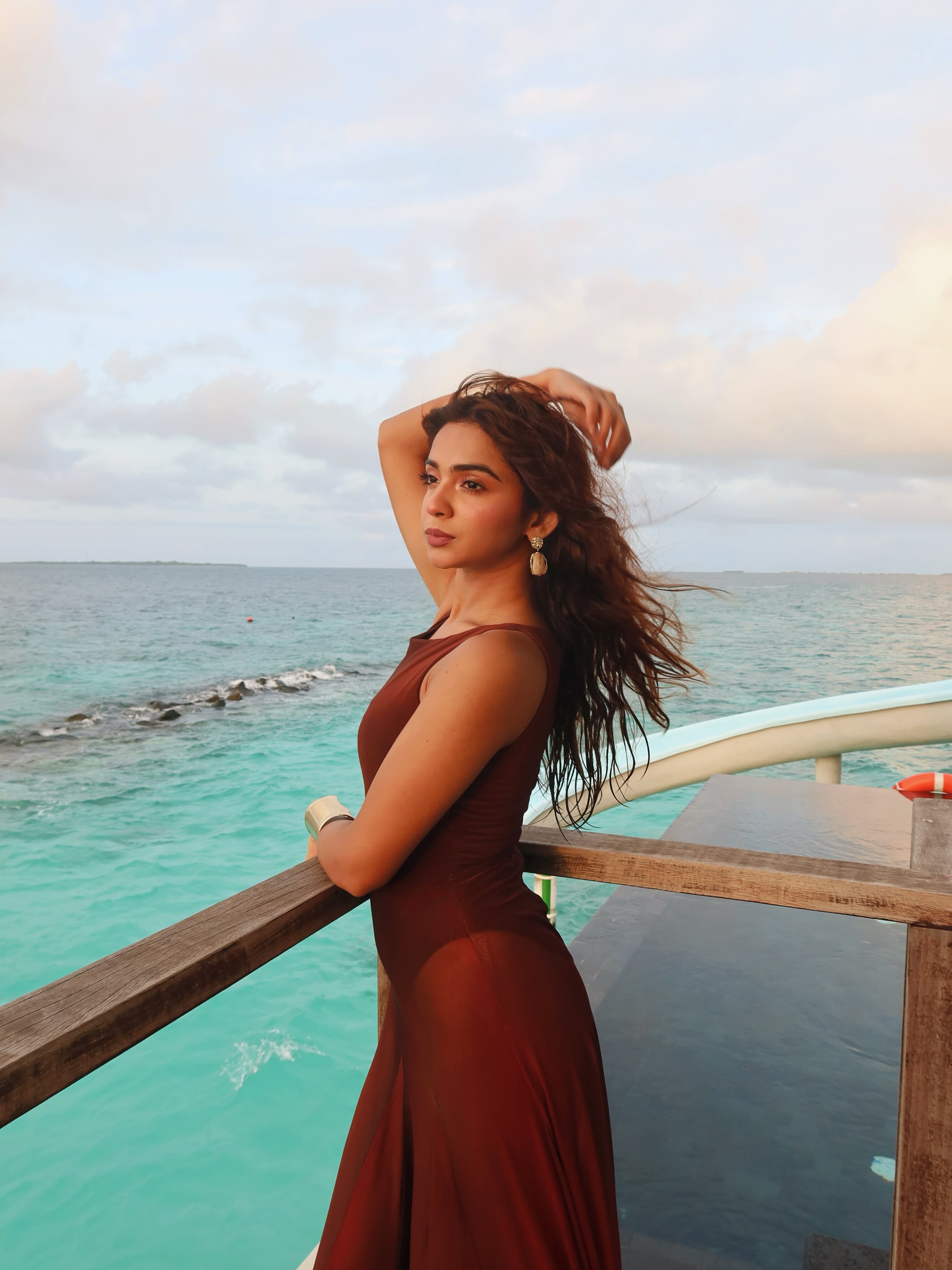 Woman in a sleeveless rust-brown maxi dress leaning on a wooden deck railing above turquoise sea under a cloudy evening sky