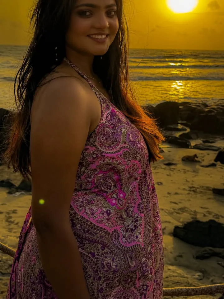 Smiling woman with long dark hair in a pink paisley halter dress on a rocky beach at sunset, with the sea behind her