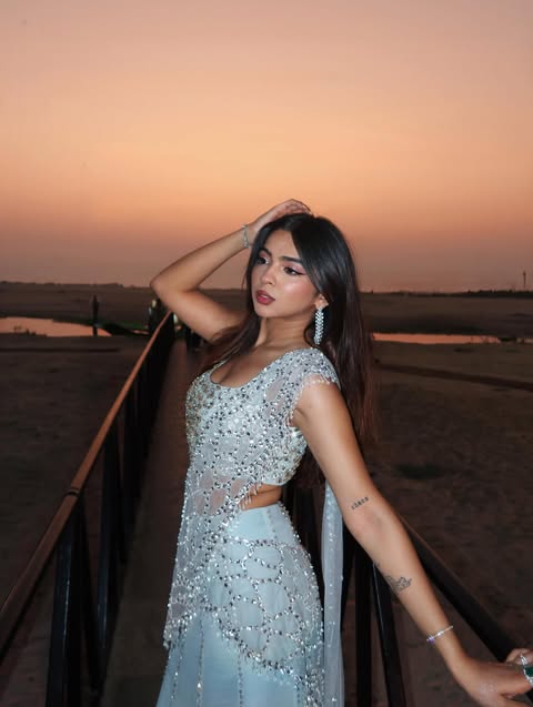 Woman with long dark hair in a sparkling pale blue embellished outfit posing by a railing at sunset