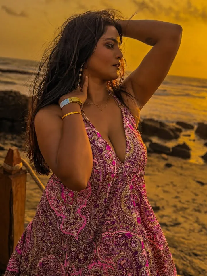 Woman with long dark hair in a pink paisley halter dress posing on a rocky beach at sunset