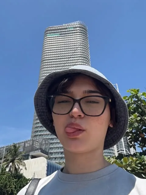 Young person in a bucket hat and black glasses sticking out their tongue in a selfie, with a tall glass tower behind
