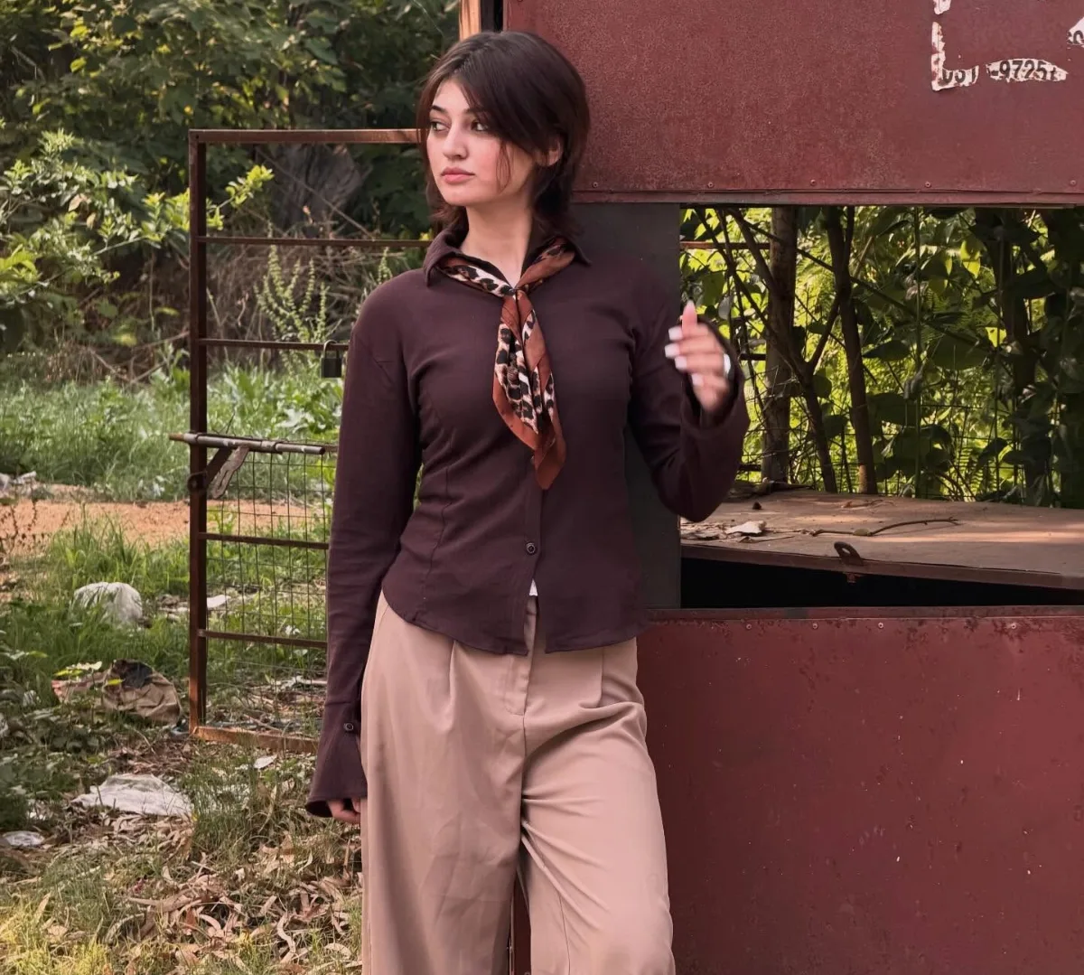 Young woman in a brown shirt, leopard-print neck scarf and beige trousers leaning on a rusty metal structure outdoors