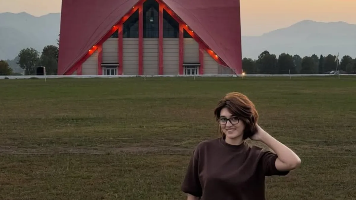imsha-rehman-news-featured Smiling woman with short brown hair and glasses stands on a grass field before a red triangular building at dusk