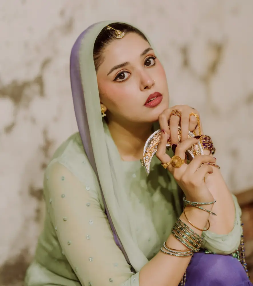 Woman in a pale green outfit and sheer headscarf with purple trim, wearing gold bangles and rings