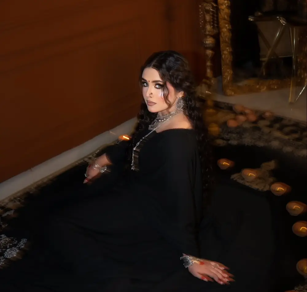 Woman with long dark curly hair in a black flowing outfit and silver jewelry, seated among glowing candles on a rug