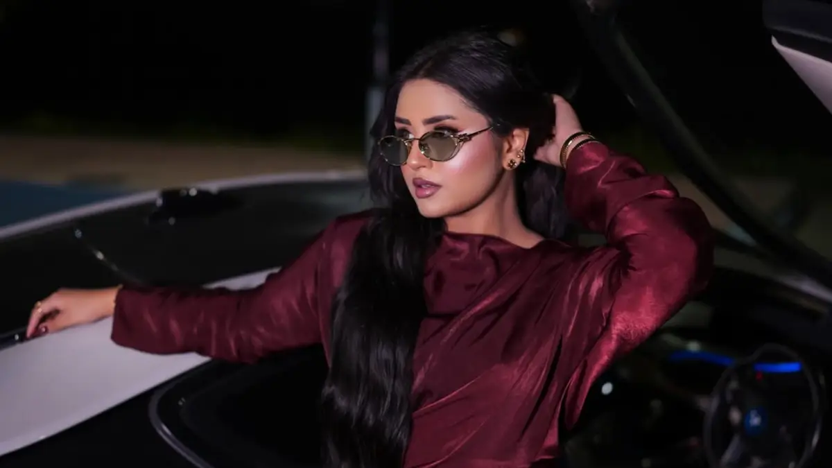 minahil-malik-featured Woman with long dark hair in tinted gold-framed sunglasses and a burgundy satin top, seated in a car at night