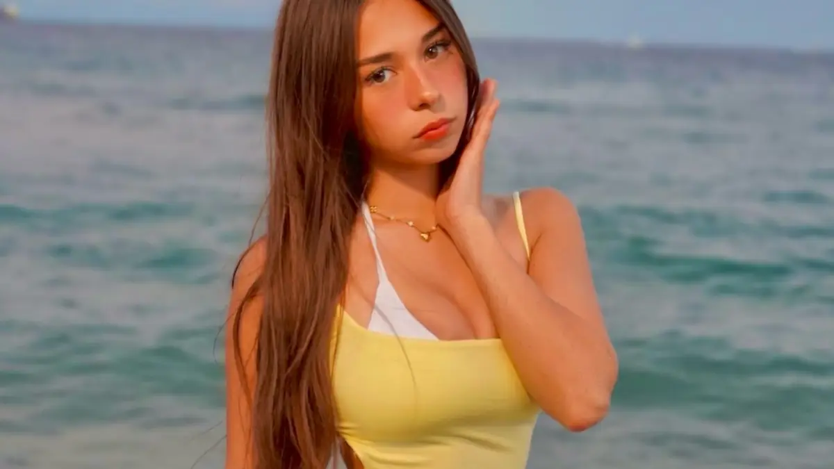 sophie-rain-featured Young woman with long brown hair in a yellow top and gold necklace, one hand touching her cheek, with the sea behind her